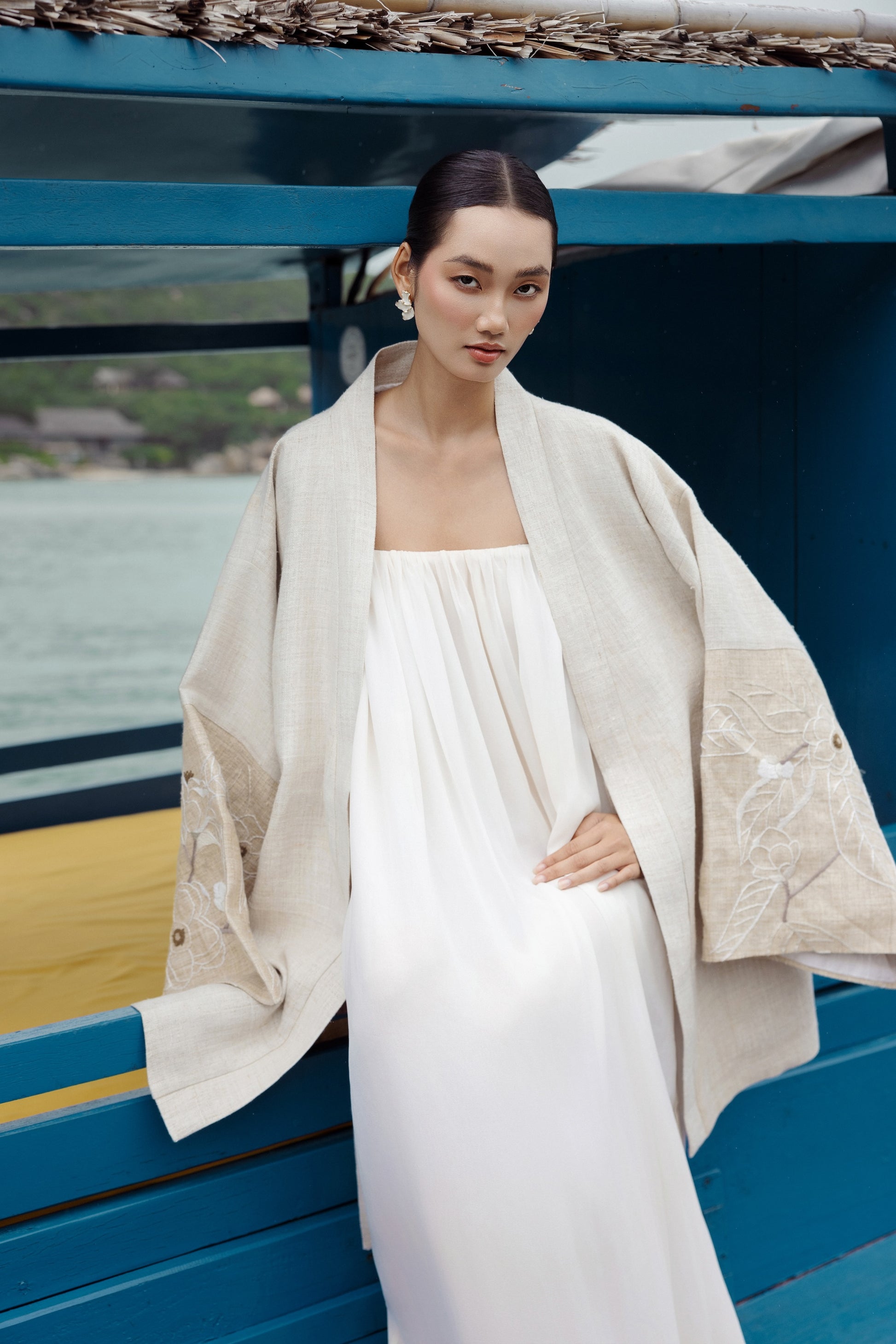 Woman in a white dress and beige coat standing on a boat with a scenic background
