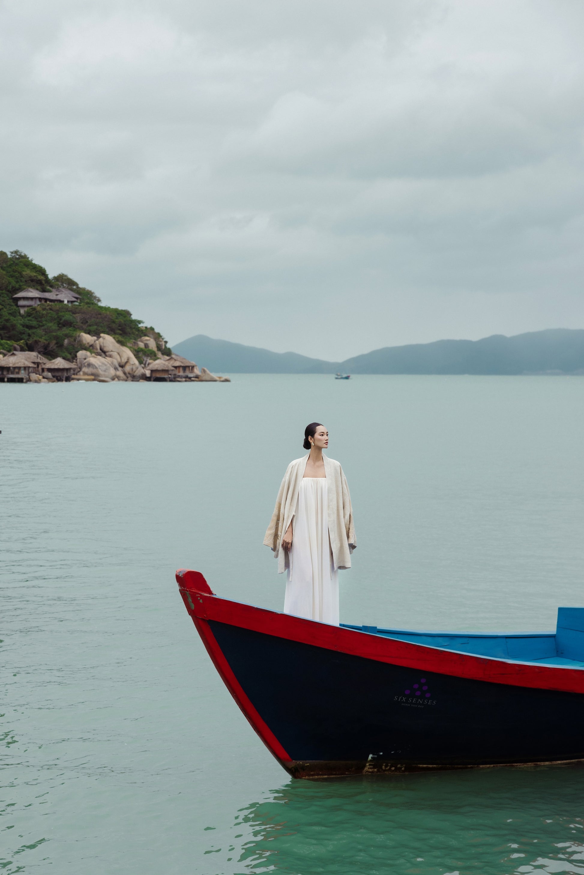 Woman in a white dress standing on a red and blue boat in a calm body of water with mountains in the background.