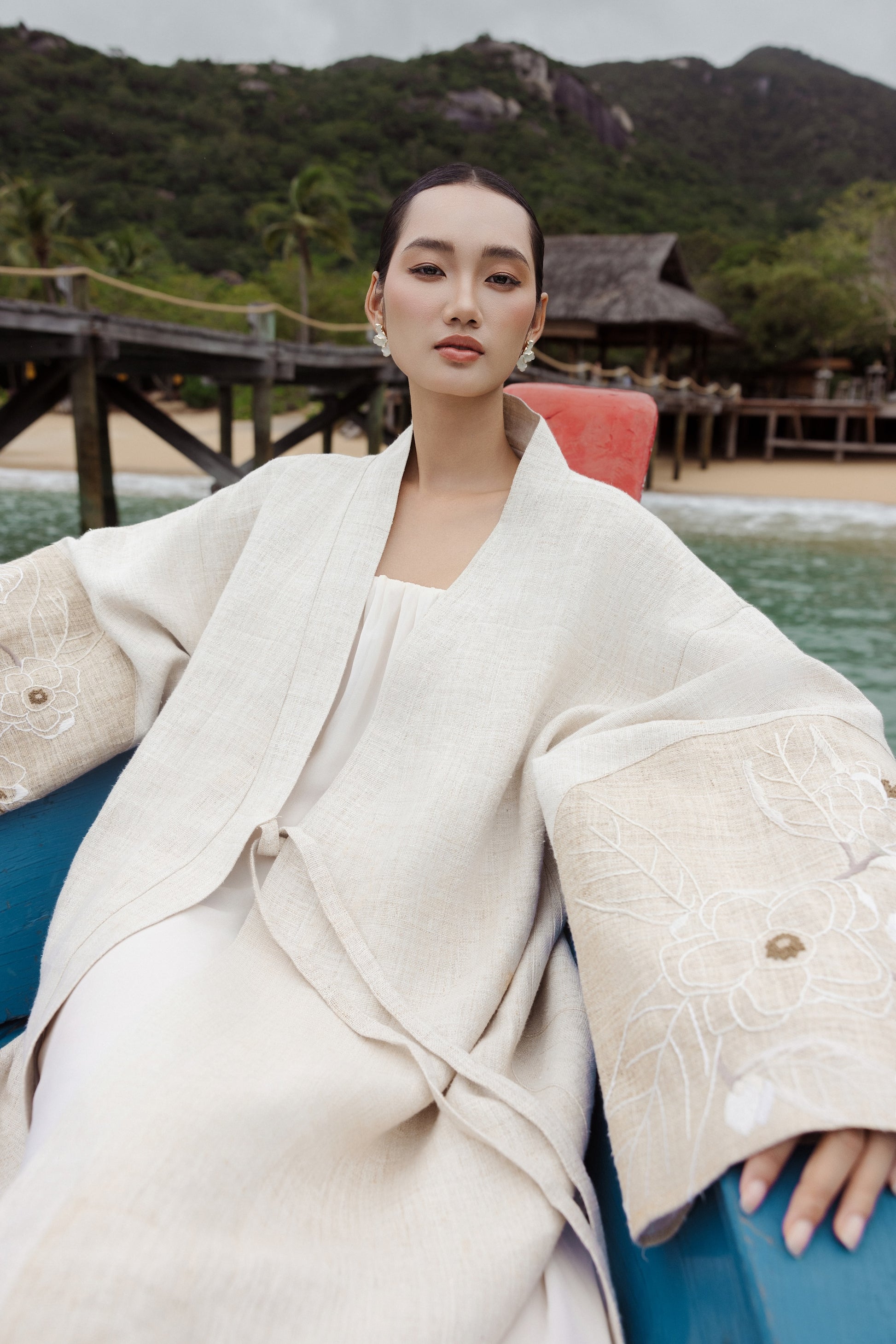Woman in a beige outfit sitting on a bow of a boat with a tropical background