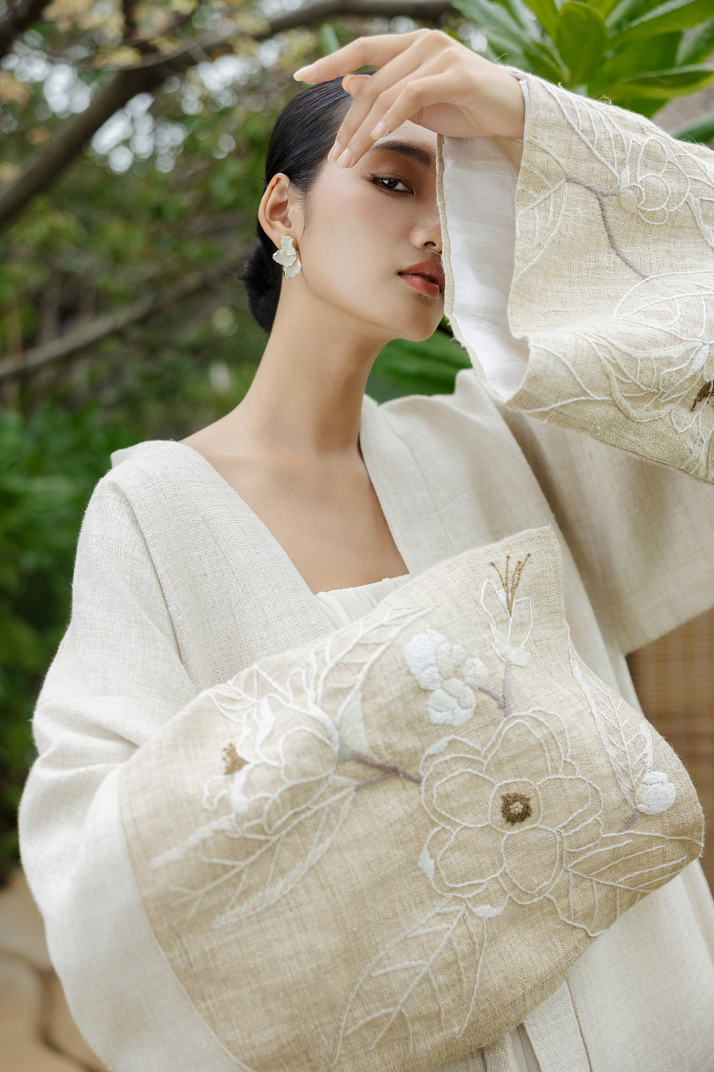 Woman in traditional white garment with floral embroidery, surrounded by greenery.