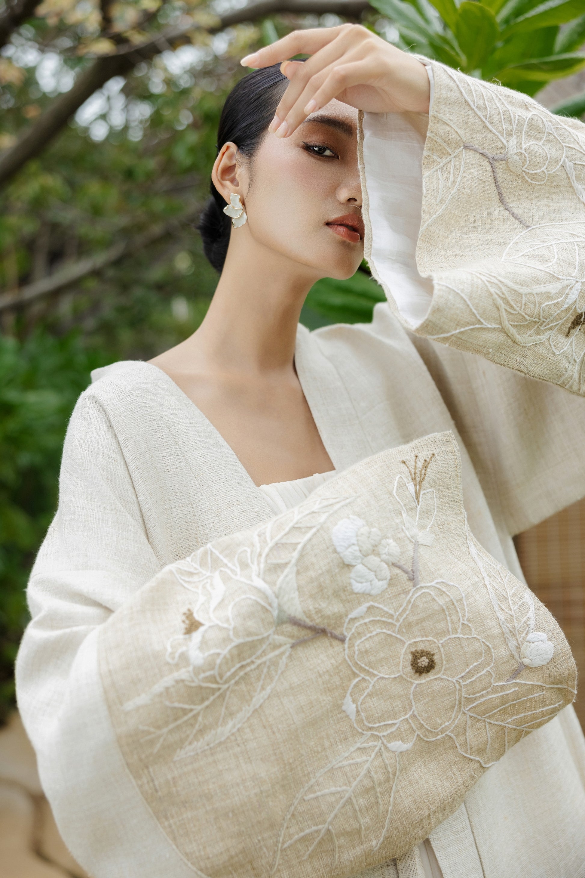Woman in traditional white garment with floral embroidery, surrounded by greenery.