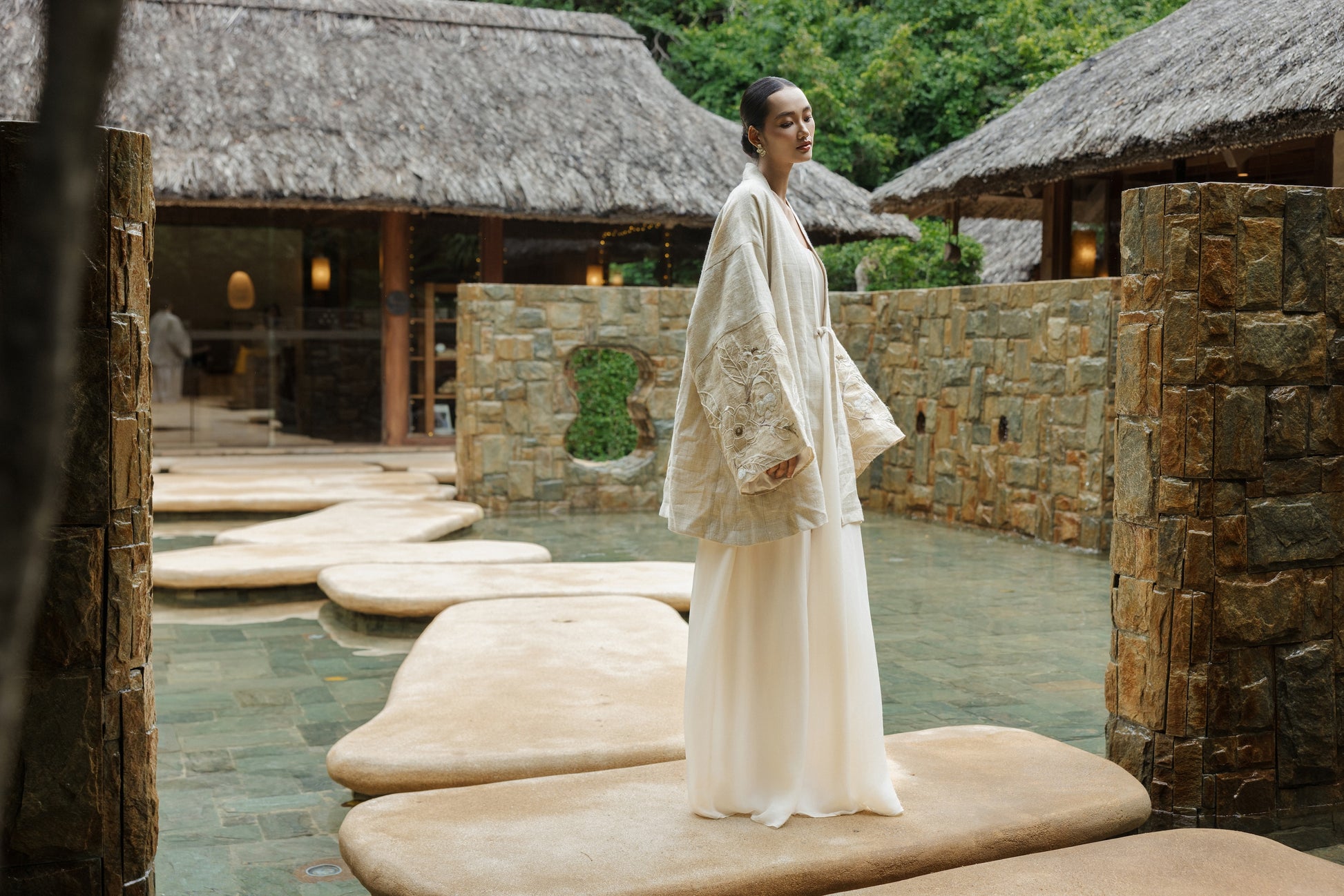 Woman in traditional attire standing on stone platforms near stone buildings with thatched roofs.